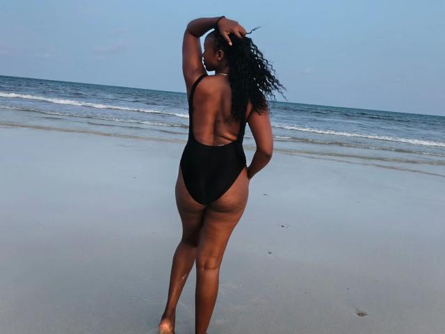 Yemaya33-hot stands on a beach, back to the camera, wearing a black thong swimsuit, her curvy ass and thick thighs prominent, long curly hair in hand.