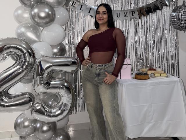 Emma Waltter, con lunghi capelli neri, indossa un top corto a spalla sola color borgogna e jeans a vita alta, ed è in piedi accanto a un palloncino argentato a forma di "25", mostrando il pollice in su con un tavolo dietro di lei..