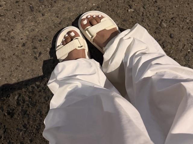 Los pies de Emma Waltter, con sandalias blancas y tiras beige, lleva pantalones blancos holgados, y está de pie sobre un terreno áspero y marrón oscuro. Sus uñas de los pies están pintadas de blanco..