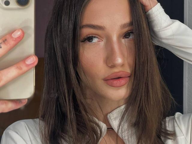 SophieKitty met lang bruin haar, roze lippen en een witte blouse, die een telefoon vasthoudt in haar linkerhand en de camera aankijkt met een subtiele, sexy uitdrukking..