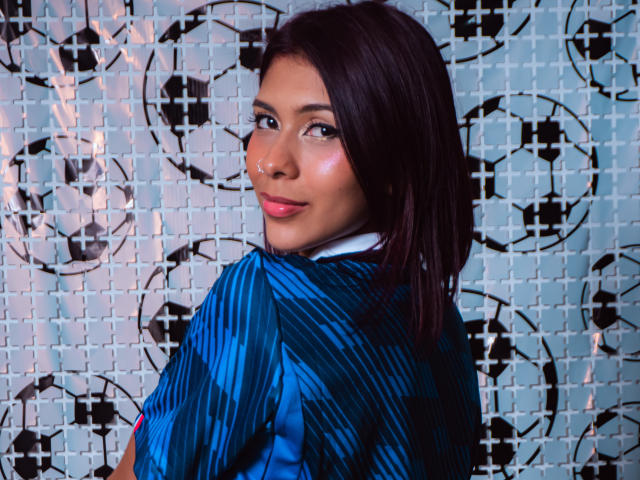Danika Serov, con i capelli castano scuro, le labbra rosate e una camicia a righe blu, guarda oltre la spalla con un leggero sorriso, mostrando un po' del suo brillante sedere..