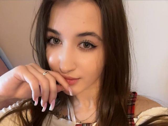 MisscharyNessy with brown hair, green eyes, and pink lips, touching her chin with a finger, wearing a plaid top, showing off her nails and ring.