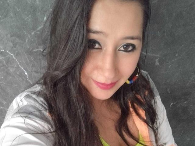 MelodyRosses has long dark hair, brown eyes, pink lipstick, and a small smile. She's wearing a colorful earring and a gray shirt with a green top visible.