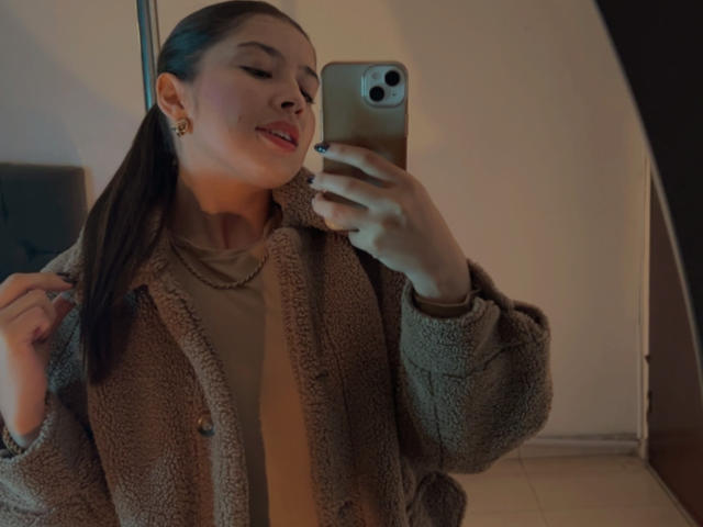 EmmaLombardi with p ponytail, wearing fuzzy brown jacket, holding gold phone, smiling, red lipstick, gold earrings, necklace, standing in dimly lit room, taking selfie.