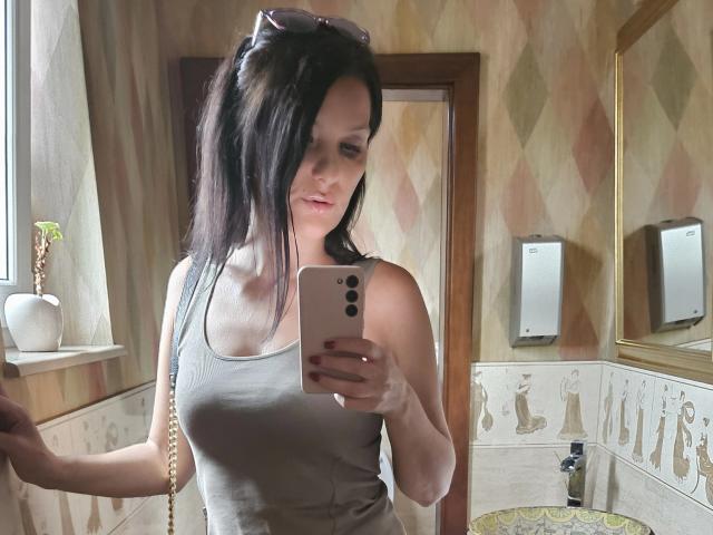 JoliDanielle with long black hair, gray tank top, holding phone, taking mirror selfie, small boobs, fair skin, pink lipstick, sunglasses on head, light brown walls, potted plant.