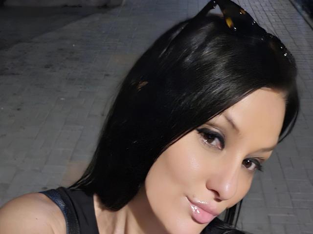 JoliDanielle has shiny black hair, brown eyes, and a slight smile. She's wearing a black top with sunglasses on her head. Her glossy lips are slightly parted.