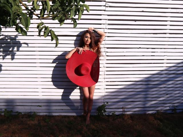 KayaBelle se tient pieds nus devant un mur en bois blanc, tenant un grand chapeau rouge avec ses seins et son bras, des feuilles vertes au-dessus, la lumière du soleil projetant des ombres..