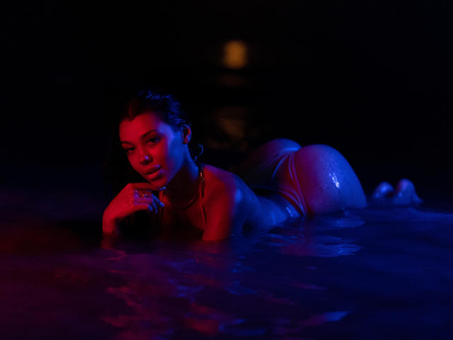 NatashaDavie lies in dark water, illuminated by red and blue lights, with wet skin and small, firm tits, wearing tight pink booty shorts, and looking seductively at the camera.