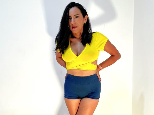 JadeFoster with black hair, yellow crop top showing her tits, and blue shorts, standing with hands on her hips, smiling at the camera. Her tits are small and perky.