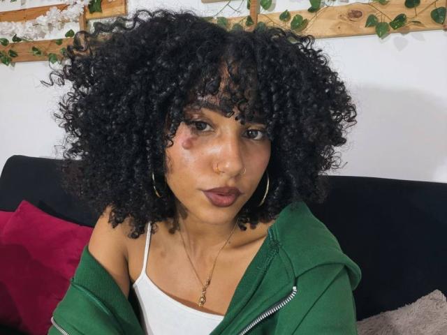 CinthiaWalker with curly black hair, medium brown skin, wearing a green zip-up jacket, white tank top, hoop earrings, and a nose ring, sitting on a black sofa.