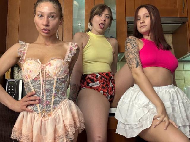 Three HotCaramelsTrio women in a kitchen: one with a pink lace corset, another with a yellow top and red patterned booty shorts, and the last with a pink crop top.