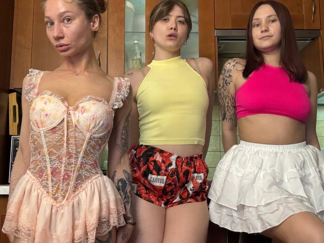 Three HotCaramelsTrio standing in a row. The left has pink lace lingerie, the middle has a yellow top and red patterned shorts, the right has a pink top and white skirt.