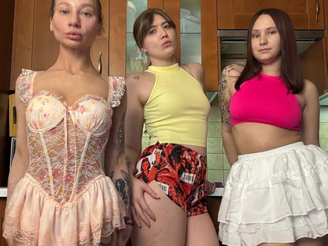 Three HotCaramelsTrio standing in a kitchen. One with pink lace lingerie, another in a yellow top and red patterned shorts, and the third in a pink top and white skirt.