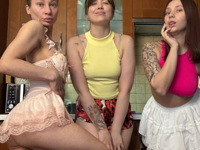 Three HotCaramelsTrio standing in a kitchen. Left: small pink lace lingerie, ass visible. Center: yellow tank, red floral shorts, tattoos. Right: pink crop top, white ruffled skirt, pouty face.