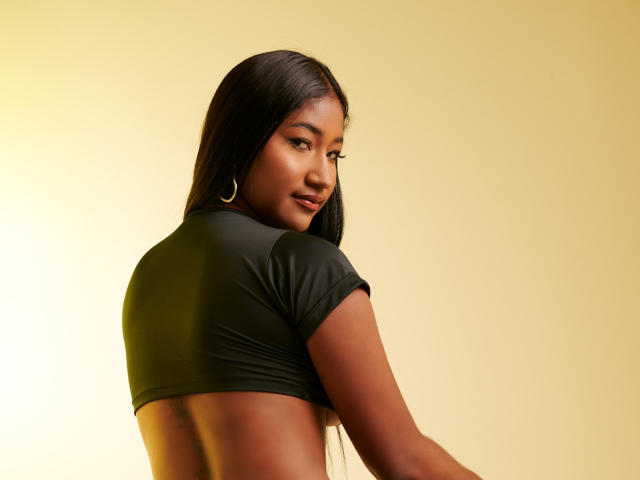 Una mujer india con cabello negro largo, piel oscura, aros grandes, mirando por encima del hombro, vestida con un top corto negro ajustado, mostrando su espalda y un poco de su trasero..