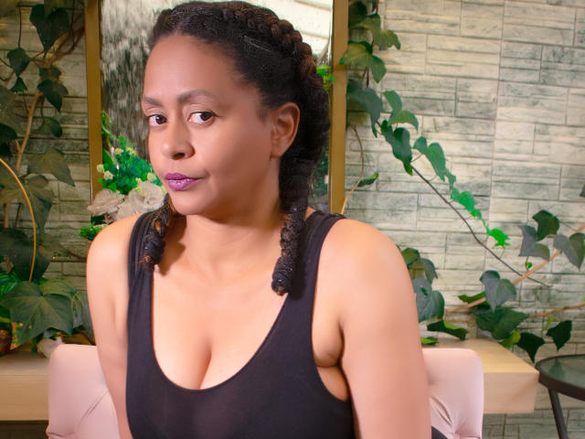 CristineHolls mit dunklen, lockigen Zöpfen, trägt ein schwarzes Tanktop, das ihre Brüste zeigt, lila Lippenstift, sitzt vor einem grünen, blättrigen Hintergrund und blickt zur Seite..