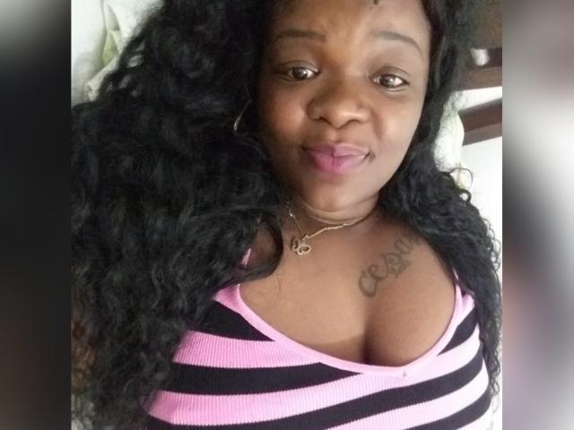 BigBbwDanay com cabelo preto encaracolado, olhos castanhos, blusa listrada rosa e preta, mostrando seus seios grandes com a tatuagem "CRAZI" no peito, sorrindo com batom rosa..