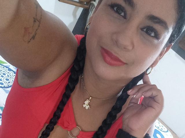 AlisonHank with braided black hair, red lipstick, hoop earrings, and a necklace with a small pendant, holding her chin, showing a tattoo on her arm, in a red top.
