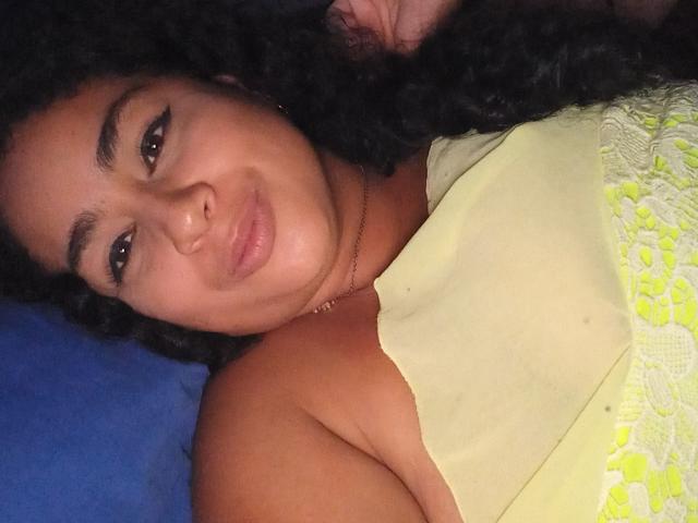 AlisonHank with curly black hair, tan skin, and eyeliner, lying on a blue surface, wearing a yellow dress with white lace, smiling at the camera.