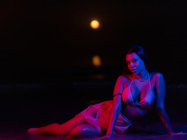 NatashaDavie is sitting on a beach at night, wearing a pink bikini, with blue and red lighting highlighting her boobs, ass, and long dark hair. A glowing orb is in the background.