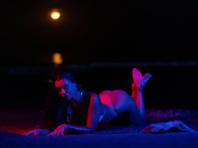NatashaDavie is lying on a dark beach at night, wearing a shiny silver bikini, with red and blue lights highlighting her ass and legs, and a glowing moon above.