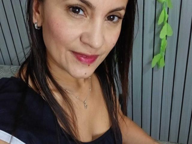 SolaraDubois has a slight smile, pink lipstick, small boob cleavage, brown eyes, straight dark hair, nose piercing, and a small chin bruise. She wears a black top and necklace.