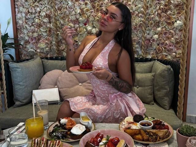 NatashaDavie with long brown hair, glasses, pink lace dress, eating cake, showing tits, tattoo on arm, surrounded by breakfast, fruit, and drinks on green sofa.