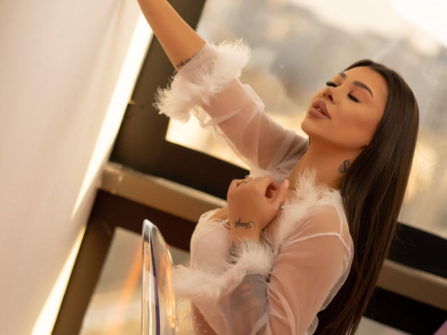 NatashaDavie with long brown hair, wearing a sheer white robe with fluffy cuffs, touching a window, showing tattoos on her hand and neck, with her eyes closed, pouty lips.
