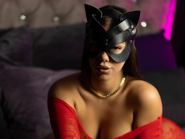 NatashaDavie with cat mask, red off-shoulder top, and gold chain, showing her boobs, dark hair, and serious face, in a dimly lit, sexy bedroom.
