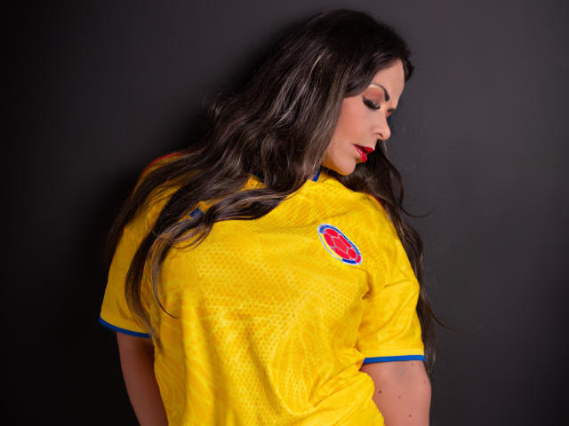 MelissaLeen with long brown hair, red lipstick, wearing a yellow sports jersey with blue trim, and a red circular logo on the back, looking down, against a dark background.
