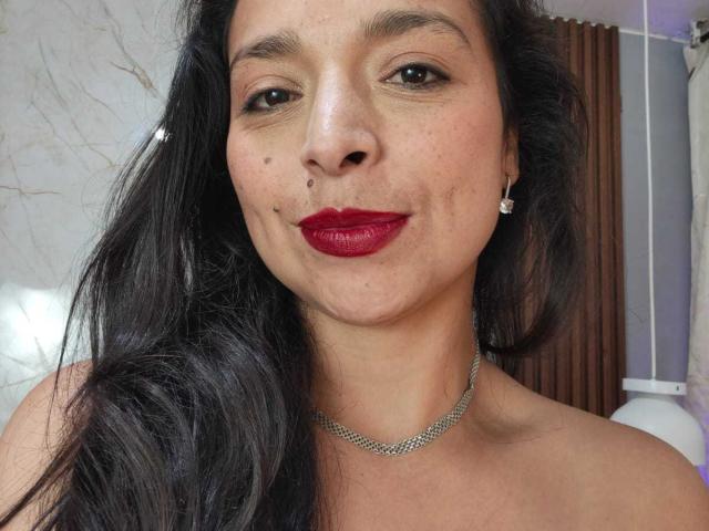 StangelarRivera has dark long hair, red lipstick, a small mole on her left cheek, and a silver chain necklace. She's wearing small earrings and smiling slightly.