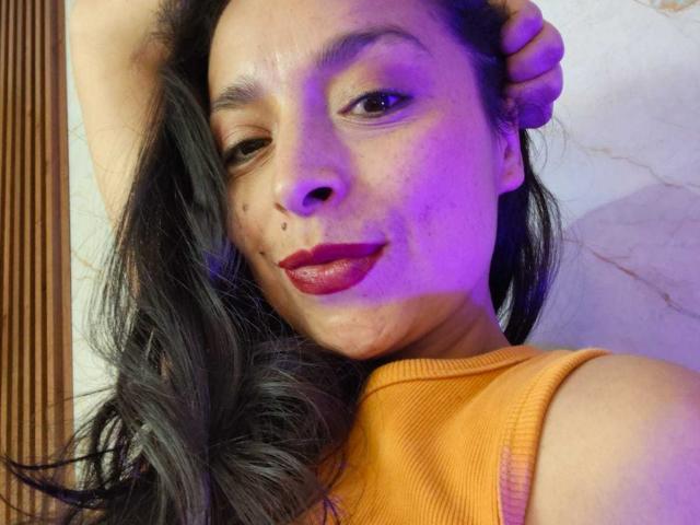 StangelarRivera with wavy black hair, red lipstick, and a cute mole on her right cheek, wearing an orange top, winking, and touching her head with a hand.