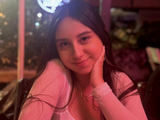 RosalieDupont with long brown hair, pink shirt, and necklace, smiling softly, resting her cheek on her hand, in a dimly lit, pink-tinged environment.