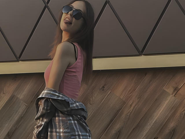 Tora Stambaugh, con cabello castaño, vestida con una camiseta rosa, gafas de sol negras y una camisa de cuadros puesta sobre sus glúteos, mirando hacia atrás con una sonrisa..