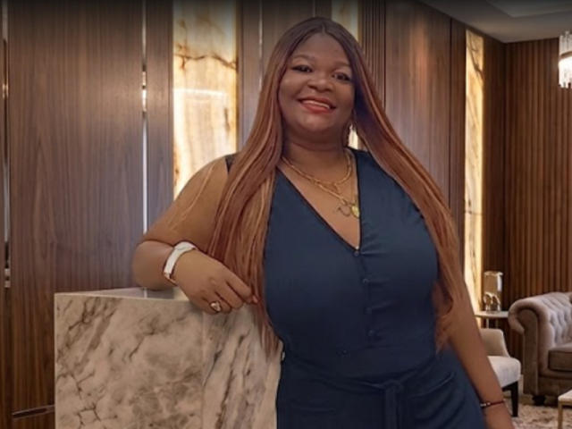BigBbwDanay met lang bruin haar, een blauwe jurk, grote borsten en een glimlach, draagt een gouden ketting en armband, leunt tegen een marmeren muur en glimlacht in een houten kamer..
