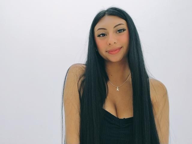 MadissonStar, con lunghi capelli neri, indossa una maglietta nera, mostra il seno, sorride, ha la pelle chiara, rossetto rosso e una piccola collana d'argento, su uno sfondo bianco uniforme..