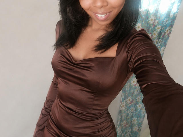 KattyBellah with dark brown, silky dress, showing her tits and smiling, her black hair falling to the sides, holding a phone for a selfie.