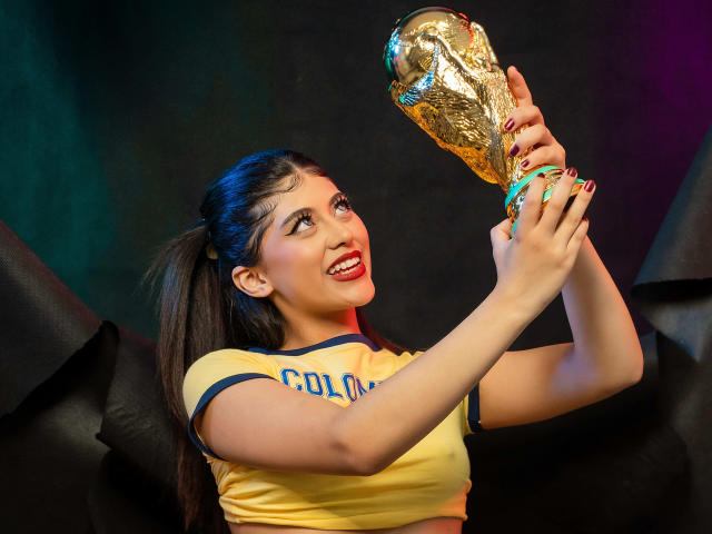 CoriinneBenson holding a golden FIFA World Cup trophy with a happy, open-mouthed smile. She's wearing a yellow crop top with blue trim, and her nails are colorful.