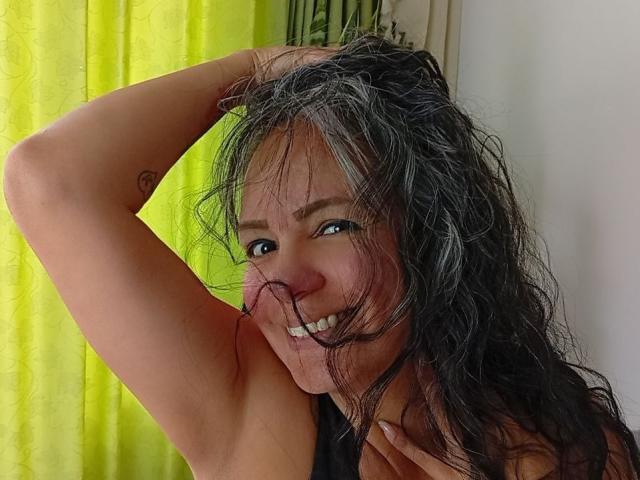 LailaCastillo with wet, messy black hair, smiling, raising her arm to show a small tattoo on her bicep, green curtain in the background. Boobs not visible, just her face.