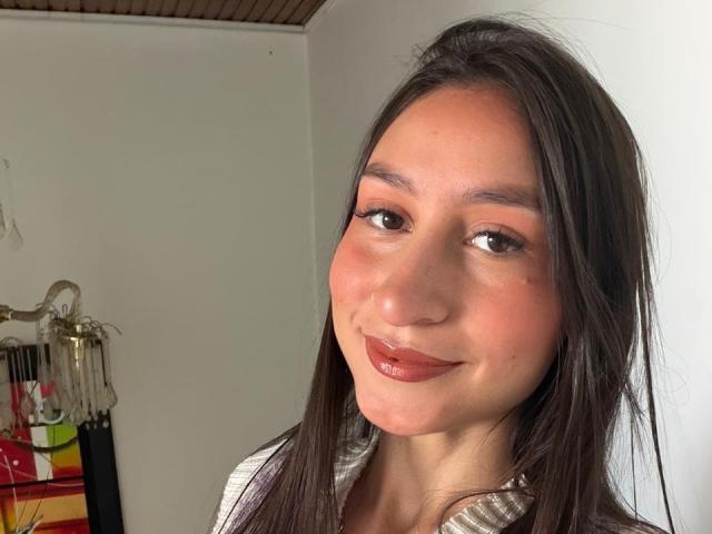 MaddyCarina with brown eyes, straight long dark hair, light tan skin, pinkish-red lips, slight smile, wearing a striped shirt, looking at camera, white wall background.