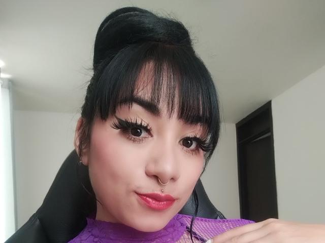 SthephanaLise ha i capelli neri, sopracciglia folte, ciglia lunghe, rossetto rosa e un piercing al naso. Indossa una maglietta viola ed è seduta su una sedia nera..