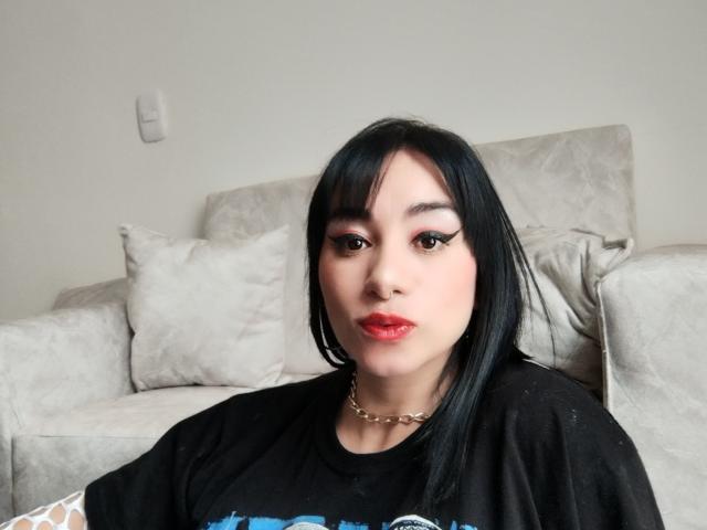 SthephanaLise with straight black hair, pink eyeshadow, red lipstick, black shirt, gold chain, sitting on a beige couch. Her tits are small, she's looking at the camera.