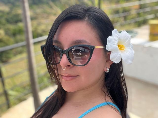 EmaJames, avec des lunettes de soleil noires, un anneau au nez et une fleur de frangipanier blanche dans ses cheveux foncés, portant un haut bleu, souriant légèrement, montrant son joli visage et son petit nez..