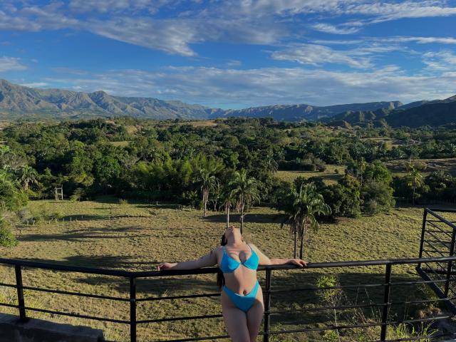EmaJames se tient les bras ouverts, vêtue d'un bikini turquoise, mettant en valeur ses grandes seins et ses petites fesses, sur un paysage ensoleillé, montagneux et verdoyant, appuyée contre une rambarde en métal noir..