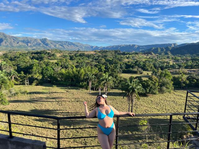 EmaJames en maillot de bain bleu, debout sur une rambarde noire, les bras levés, montrant ses seins, avec un paysage de montagne et un ciel dégagé en arrière-plan..