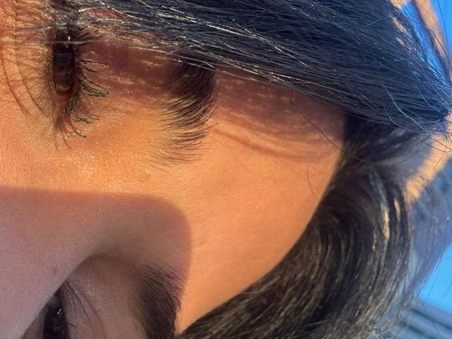 L'œil et la joue supérieure de MajoJones, avec ses cils noirs, sa paupière marron et ses courts cheveux bruns, baignés de lumière vive. Sa peau est légèrement brillante, révélant sa texture naturelle..