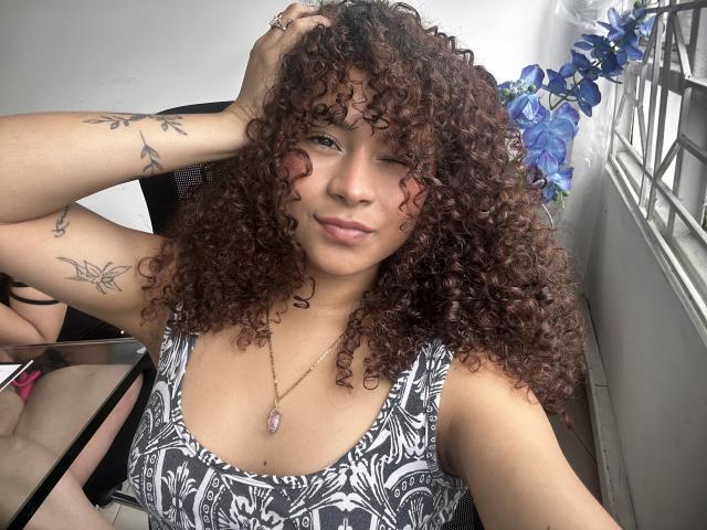 SofiaPassionne tem cabelos castanhos cacheados, seios médios, tatuagens nos braços, veste um vestido com padrão preto e branco e um colar rosa, piscando para a câmera..