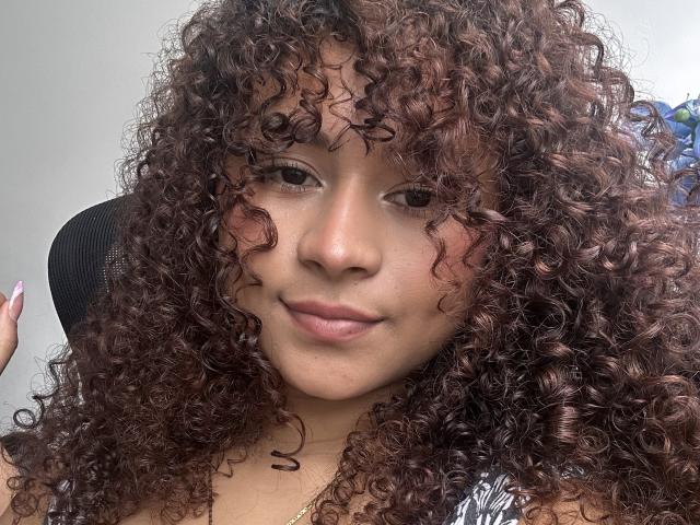 SofiaPassionne tiene el cabello castaño rizado, piel de color marrón claro, y está sonriendo suavemente con lápiz labial rosa. Su mano derecha es ligeramente visible y tiene las uñas pintadas de rosa..