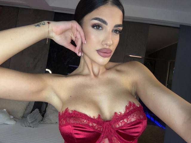 RaisaKali con un sujetador de encaje rojo, mostrando sus grandes senos, cabello oscuro y un tatuaje en la muñeca, sonriendo a la cámara en una habitación bien iluminada..