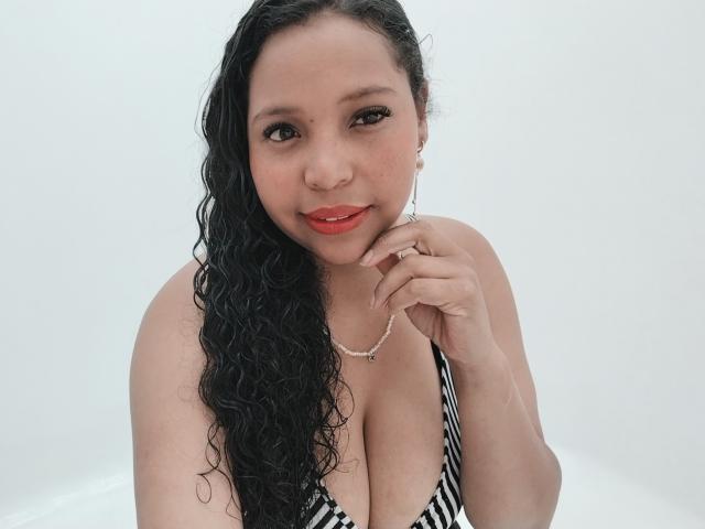 Sthefanni tiene el cabello negro y rizado, usa lápiz labial rojo y lleva un bikini a rayas que muestra un busto grande. Está sosteniendo su barbilla con un collar de plata..